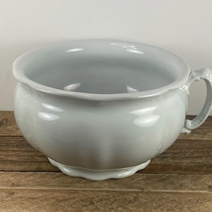 May include: A light gray, antique chamber pot with a handle. The ceramic pot has a wide, scalloped rim and a decorative base. The handle is curved and attached to the side of the pot. The pot is sitting on a wooden surface.
