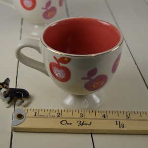 May include: A white ceramic teacup with a red and pink apple design. The inside of the cup is a solid red color. The cup has a gold rim and a small foot.