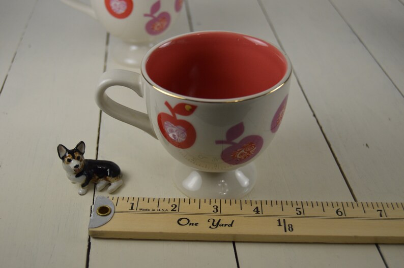 May include: A white ceramic teacup with a red and pink apple design. The inside of the cup is a solid red color. The cup has a gold rim and a small foot.