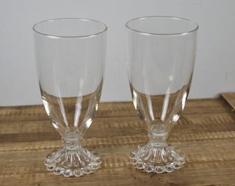 May include: A pair of clear glass footed tumblers featuring a smooth, cylindrical upper bowl that transitions into a decorative, fluted base adorned with a ring of small, raised glass beads around the bottom edge of the pedestal stand.
