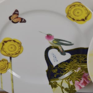 Puede incluir: Dos platos de cer&aacute;mica blanca con dise&ntilde;os decorativos. Un plato presenta un colibr&iacute;, una mariposa y flores amarillas. El otro plato tiene un dise&ntilde;o floral con acentos dorados.