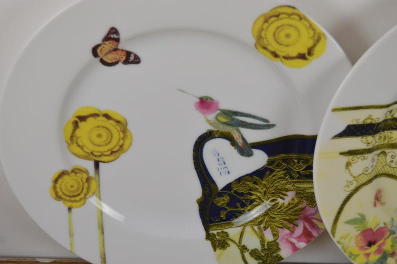 Puede incluir: Dos platos de cer&aacute;mica blanca con dise&ntilde;os decorativos. Un plato presenta un colibr&iacute;, una mariposa y flores amarillas. El otro plato tiene un dise&ntilde;o floral con acentos dorados.