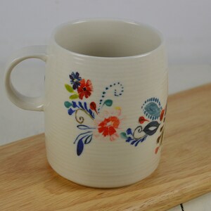 K&ouml;nnte beinhalten: Wei&szlig;e Keramiktasse mit einem Blumendesign mit blauen, roten und gr&uuml;nen Blumen. Die Tasse hat einen Henkel und steht auf einer Holzoberfl&auml;che.
