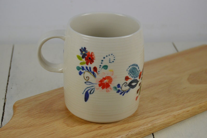 K&ouml;nnte beinhalten: Wei&szlig;e Keramiktasse mit einem Blumendesign mit blauen, roten und gr&uuml;nen Blumen. Die Tasse hat einen Henkel und steht auf einer Holzoberfl&auml;che.