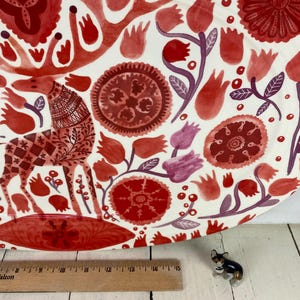 Op de afbeelding: Een grote, ronde decoratieve schaal met een witte achtergrond en een rood bloemen- en dierenontwerp. De schaal heeft een gestileerde giraffe en verschillende bloemelementen in tinten rood en paars. Een houten liniaal staat op de voorgrond.