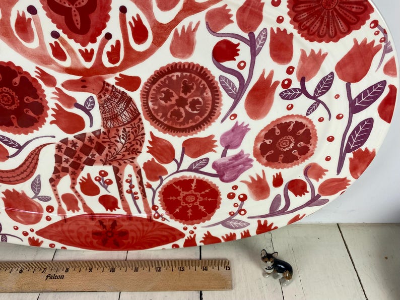 Op de afbeelding: Een grote, ronde decoratieve schaal met een witte achtergrond en een rood bloemen- en dierenontwerp. De schaal heeft een gestileerde giraffe en verschillende bloemelementen in tinten rood en paars. Een houten liniaal staat op de voorgrond.