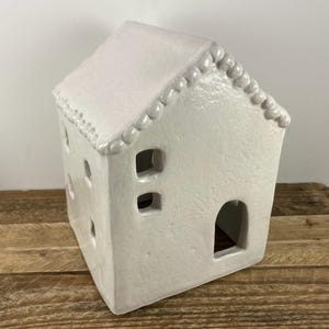 May include: A white ceramic house-shaped candle holder with a textured surface. The house has a pitched roof, small rectangular windows, and a rounded doorway. The roofline is decorated with small, rounded details. The candle holder sits on a wooden surface.