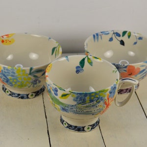 May include: Three ceramic bowls with a floral design. The bowls are white with blue, yellow, green, and orange flowers. The bowls have a pedestal base.