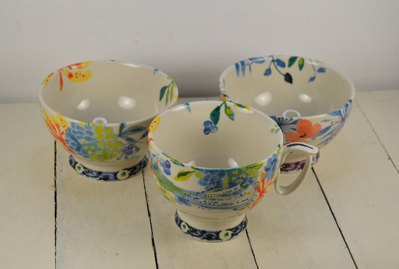May include: Three ceramic bowls with a floral design. The bowls are white with blue, yellow, green, and orange flowers. The bowls have a pedestal base.