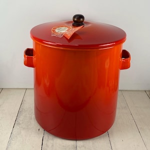May include: A large, vibrant orange enamel pot with a matching lid and wooden knob. The pot has two side handles and a glossy finish. A tag hangs from the lid. The pot sits on a white wooden surface.