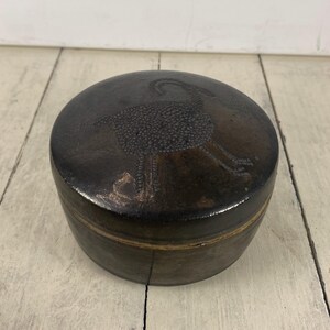 May include: A round, dark brown ceramic box with a lid. The lid features a detailed illustration of a bird. The box has a glossy finish and a band of lighter brown around the middle.