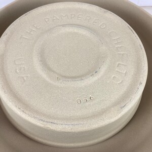 May include: Tan stoneware baking dish with a lid. The lid is embossed with "THE PAMPERED CHEF LTD" and "USA". The number "056" is stamped on the lid. The dish is sitting inside a larger, matching stoneware bowl.