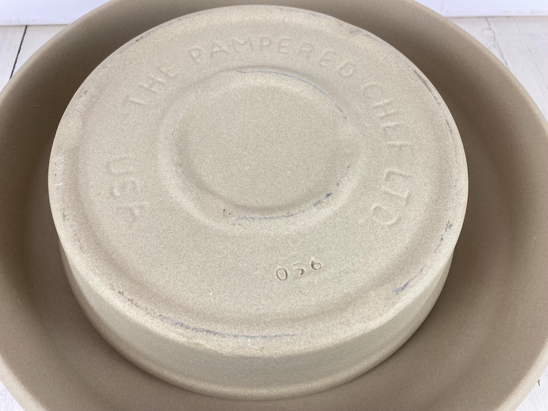 May include: Tan stoneware baking dish with a lid. The lid is embossed with "THE PAMPERED CHEF LTD" and "USA". The number "056" is stamped on the lid. The dish is sitting inside a larger, matching stoneware bowl.
