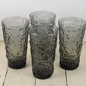 May include: Set of four gray glass drinking glasses with a textured surface. The glasses are tall and cylindrical.