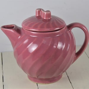 May include: A pink ceramic teapot with a swirl design and a lid with a knob on top.