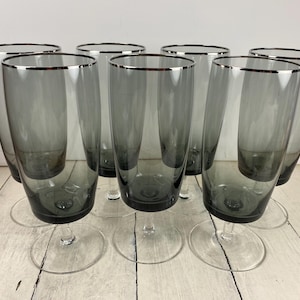 May include: A set of eight gray champagne flutes with a silver rim. The glasses have a tall, slender design and sit on clear, round bases. The stemware is arranged on a light-colored wooden surface.