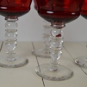 May include: Four red wine glasses with clear glass stems featuring a beaded design. The glasses are on a white wooden surface.