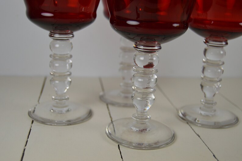 May include: Four red wine glasses with clear glass stems featuring a beaded design. The glasses are on a white wooden surface.