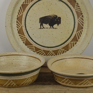 May include: A set of three ceramic bowls with a speckled beige glaze and brown and green geometric designs. The largest bowl features a silhouette of a buffalo in the center.