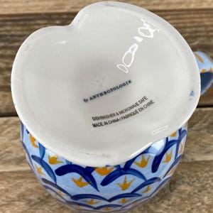 Op de afbeelding: Een witte, hartvormige keramische creamer met een blauw en geel bloemmotief. De creamer is gemarkeerd met "by Anthropologie" en is vaatwasser- en magnetronbestendig.