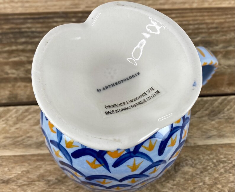 Op de afbeelding: Een witte, hartvormige keramische creamer met een blauw en geel bloemmotief. De creamer is gemarkeerd met "by Anthropologie" en is vaatwasser- en magnetronbestendig.