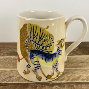 K&ouml;nnte beinhalten: Cremefarbene Keramik-Tasse mit einer farbenfrohen Illustration eines Tigers und eines gro&szlig;en, stilisierten Blattes. Der Tiger hat blaue, gelbe und violette Streifen. Die Tasse hat einen gebogenen Henkel und einen wei&szlig;en Rand.