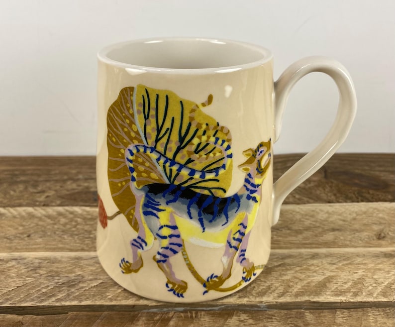 K&ouml;nnte beinhalten: Cremefarbene Keramik-Tasse mit einer farbenfrohen Illustration eines Tigers und eines gro&szlig;en, stilisierten Blattes. Der Tiger hat blaue, gelbe und violette Streifen. Die Tasse hat einen gebogenen Henkel und einen wei&szlig;en Rand.
