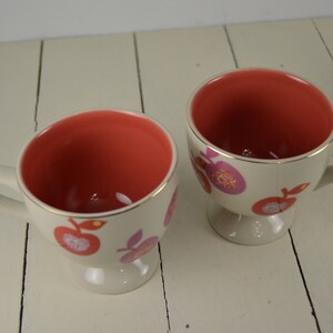 May include: Two white ceramic mugs with a pink and orange fruit design. The mugs have a gold rim and a footed base.
