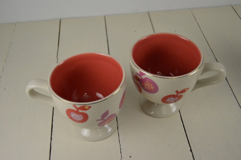 May include: Two white ceramic mugs with a pink and orange fruit design. The mugs have a gold rim and a footed base.