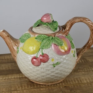 May include: A white ceramic teapot with a brown handle and a lid decorated with a strawberry, lemon, peach, and cherries. The teapot is decorated with a woven basket pattern.