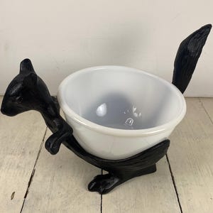 May include: A white glass bowl held by a black cast metal squirrel figurine. The squirrel features a textured surface, a raised tail, and front paws resting against the side of the bowl. The bowl is smooth, round, and sits securely within the base.