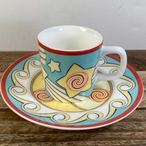 May include: A ceramic espresso cup and matching saucer set featuring a light blue background with yellow star motifs, red spiral accents, and white wavy lines. The rim of both the cup and the saucer is outlined in a thin, vibrant red border.