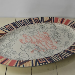 May include: An oval ceramic serving platter with a white background and a colorful geometric border. The center of the platter features a floral design with black outlines and pink accents.