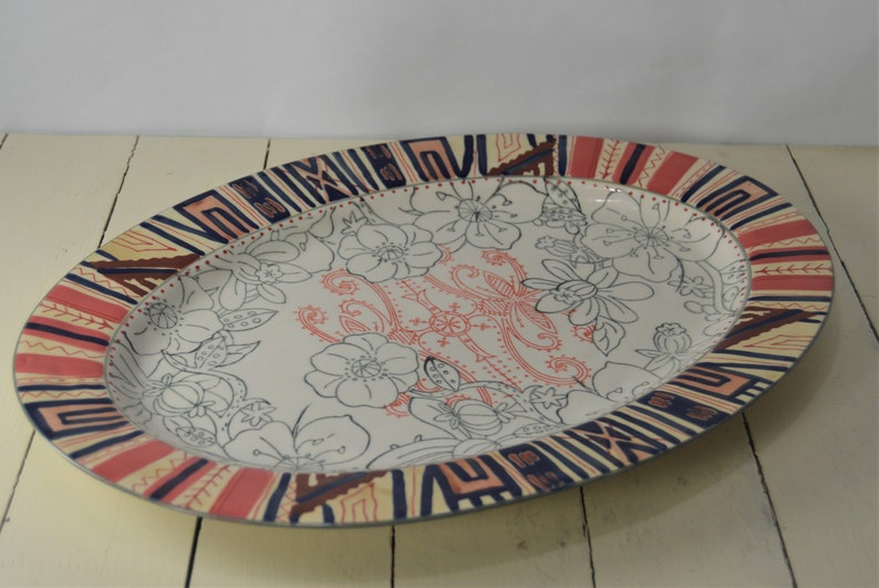 May include: An oval ceramic serving platter with a white background and a colorful geometric border. The center of the platter features a floral design with black outlines and pink accents.