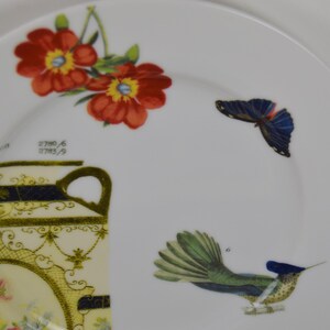 Puede incluir: Un plato de cer&aacute;mica blanco con un dise&ntilde;o floral con flores rojas y amarillas, una mariposa azul y un colibr&iacute; verde y azul. El plato tambi&eacute;n presenta un dise&ntilde;o dorado y negro con el texto "2700/6 2703/9".