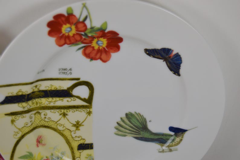 Puede incluir: Un plato de cer&aacute;mica blanco con un dise&ntilde;o floral con flores rojas y amarillas, una mariposa azul y un colibr&iacute; verde y azul. El plato tambi&eacute;n presenta un dise&ntilde;o dorado y negro con el texto "2700/6 2703/9".