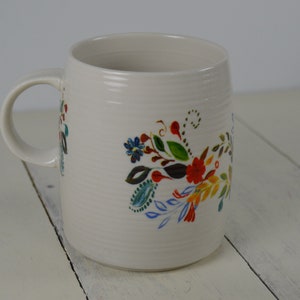 May include: Cream-colored ceramic mug with a curved handle. The mug is decorated with a colorful floral design featuring red, blue, and green flowers and leaves. The mug has a textured surface.