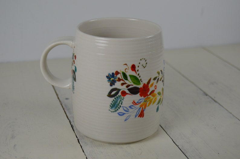May include: Cream-colored ceramic mug with a curved handle. The mug is decorated with a colorful floral design featuring red, blue, and green flowers and leaves. The mug has a textured surface.