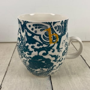 K&ouml;nnte beinhalten: Wei&szlig;er Keramikbecher mit einem t&uuml;rkis-wei&szlig;en Blumenmuster und einem gelben Buchstaben "B". Der Becher hat eine abgerundete Form mit einem wei&szlig;en Henkel.