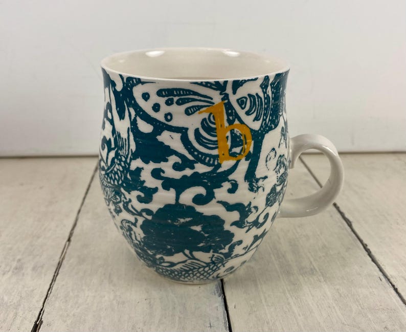 K&ouml;nnte beinhalten: Wei&szlig;er Keramikbecher mit einem t&uuml;rkis-wei&szlig;en Blumenmuster und einem gelben Buchstaben "B". Der Becher hat eine abgerundete Form mit einem wei&szlig;en Henkel.