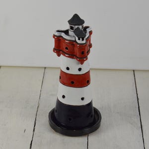 May include: A ceramic lighthouse candle holder with red, white, and blue stripes. The lighthouse has a small black roof and a white base.