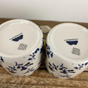 May include: Two white ceramic mugs with a blue floral design and the text "by ANTHROPOLOGIE" and a butterfly logo on the bottom. The mugs are dishwasher and microwave safe.