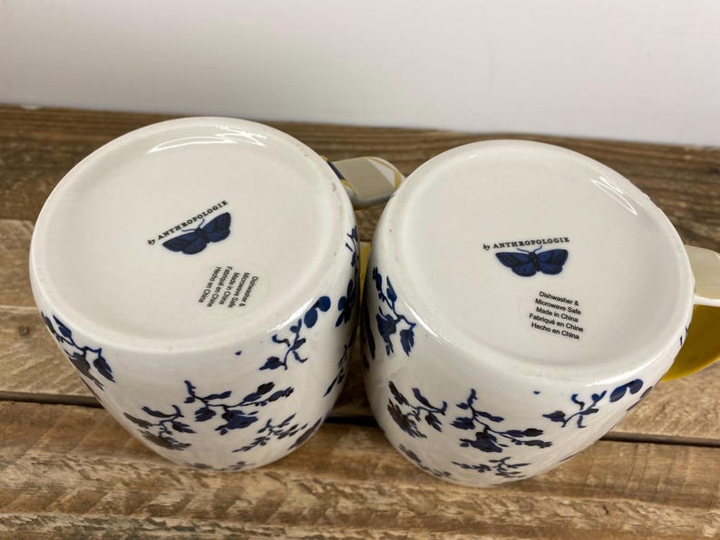 May include: Two white ceramic mugs with a blue floral design and the text "by ANTHROPOLOGIE" and a butterfly logo on the bottom. The mugs are dishwasher and microwave safe.