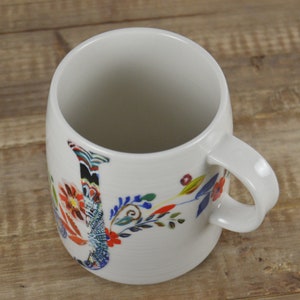 May include: White ceramic mug with a floral design. The mug features a large, colorful letter "J" with a variety of flowers and leaves in red, blue, and yellow. The mug has a curved handle and a smooth, glossy finish.
