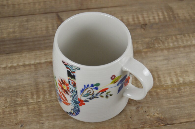 May include: White ceramic mug with a floral design. The mug features a large, colorful letter "J" with a variety of flowers and leaves in red, blue, and yellow. The mug has a curved handle and a smooth, glossy finish.