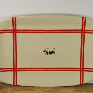 May include: Cream-colored rectangular serving platter with a red grid pattern and a black pig illustration in the center. The platter is likely ceramic or porcelain, suitable for serving food.