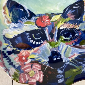 May include: A decorative ceramic plate featuring a colorful raccoon design. The raccoon's face is painted with shades of blue, green, and pink, surrounded by floral and leaf accents. The plate has a scalloped edge with a gold trim.