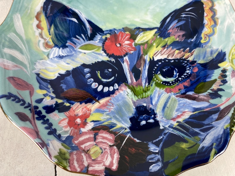 May include: A decorative ceramic plate featuring a colorful raccoon design. The raccoon's face is painted with shades of blue, green, and pink, surrounded by floral and leaf accents. The plate has a scalloped edge with a gold trim.