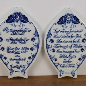 May include: Two blue and white ceramic fish-shaped plaques with Danish proverbs. The left plaque reads "At smörrebrod er ikke mad, og hierlighed er ikke had. Jeter for tiden hvad jeg ved om smörrebrod og hierlighed." The right plaque reads "Lev vel og fornoiet! Hav stedse for Diet, Du ei er ret frisk. For meget af Kiodet Saa mangen har dodet, Spis derfor helst Fisk."
