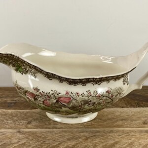 May include: Cream-colored gravy boat with a decorative rim and handle. The boat features a landscape scene with pink and green accents. The gravy boat is made of ceramic and sits on a round base.